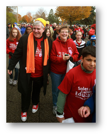 Archbishop Jerome Listecki (wearing a cheesehead zucchetto with a read walk T-shirt and Chuck Taylor high-tops) is pictured walking with NDMS fifth grade teacher and School Sister of Notre Dame Judy Fleisner. 